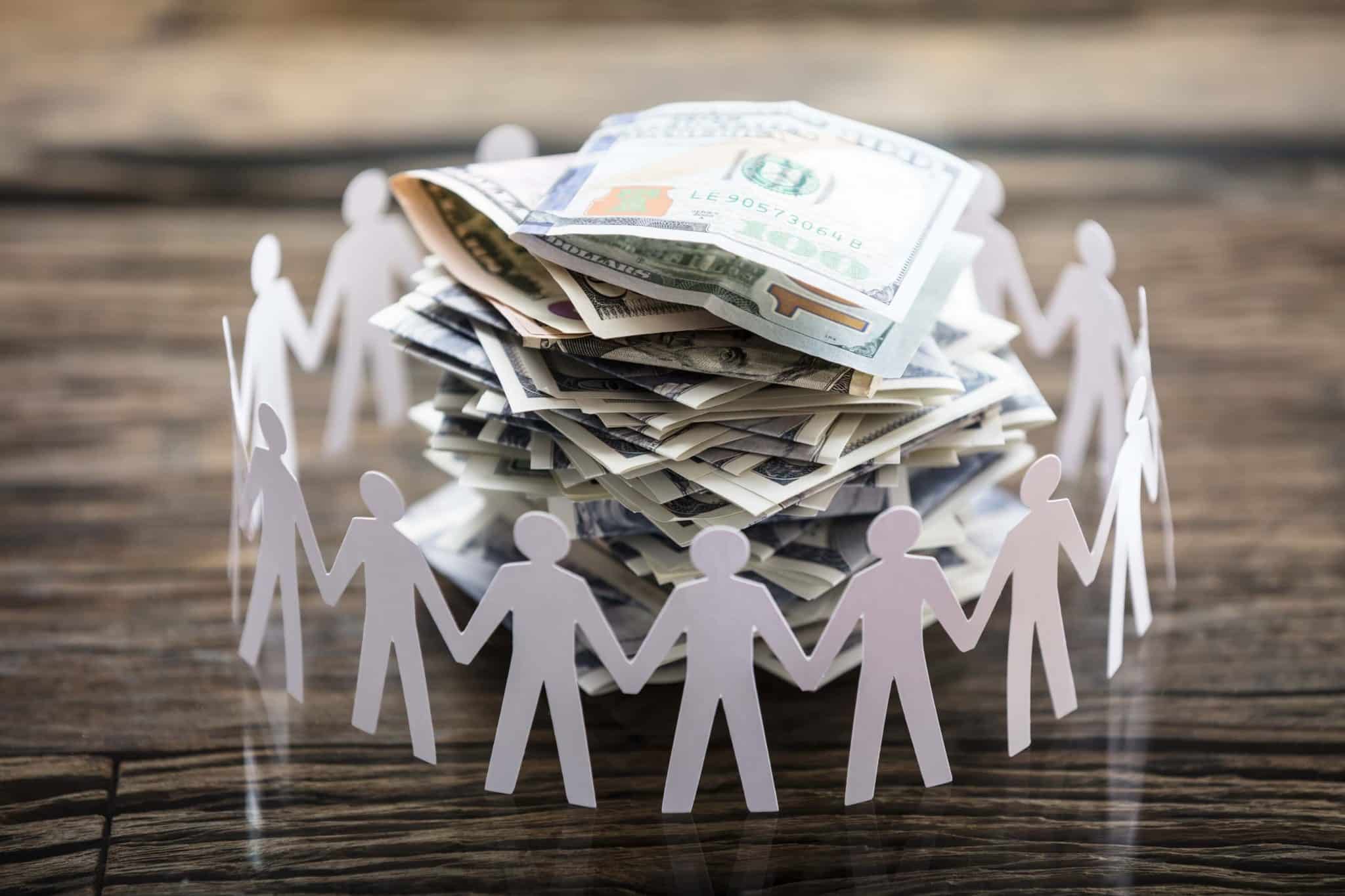 A group of paper people standing in a circle on a wooden table, surrounded by money.