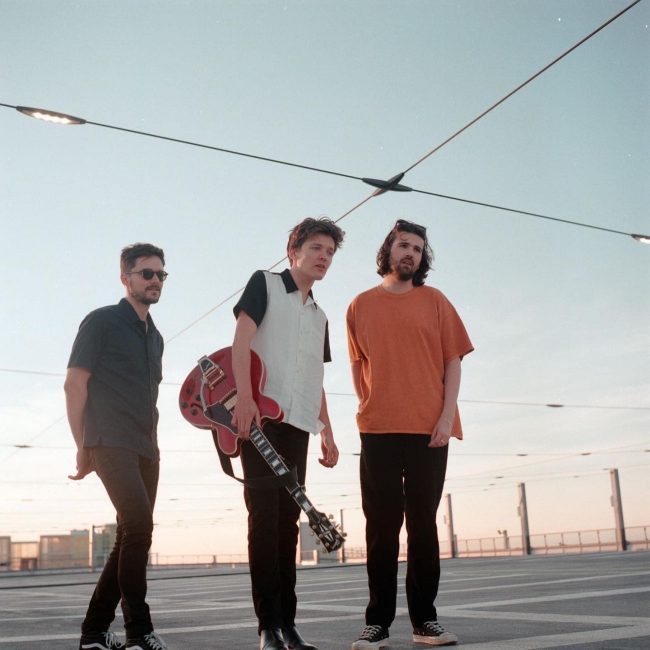 Three male members of Cusp, with the centre member holding a guitar, as they're posing against a sky backdrop